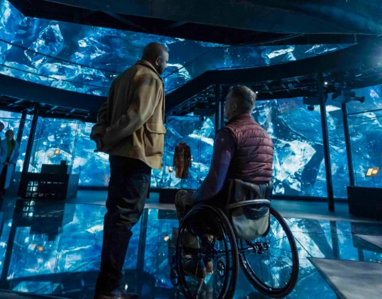 Two individuals in an immersive, blue-lit exhibition space with reflective floor and displays.