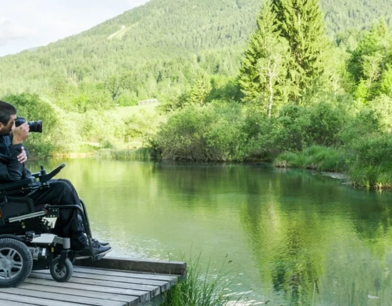 Man in wheelchair taking a picture on a wooden pier by a lake, surrounded by greenery and hills.