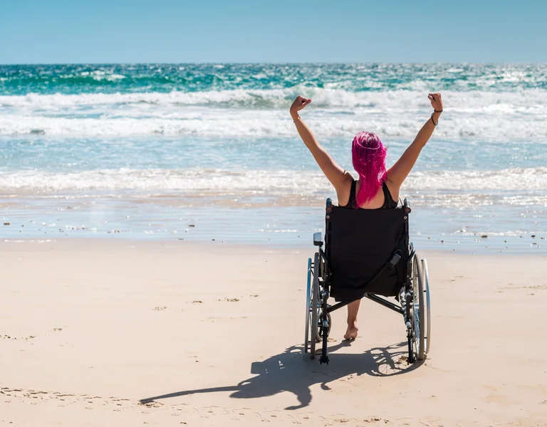 A wheelchair user is stretching her arms in front of a blue, pristine water sea on a sandy beach.