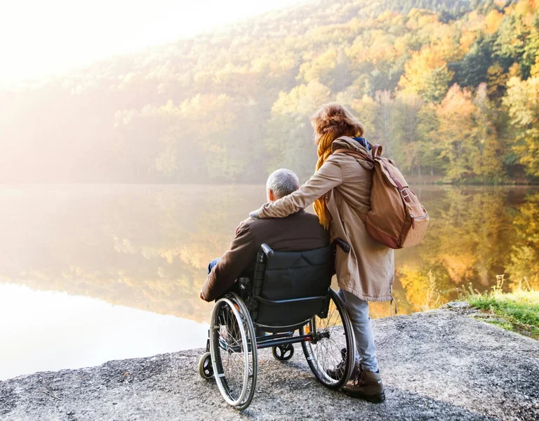 A scenic autumn view of a tranquil lake surrounded by forested hills with golden foliage. In the foreground, a person in a wheelchair sits beside a companion who has their arm around them, both enjoying the peaceful, natural setting.