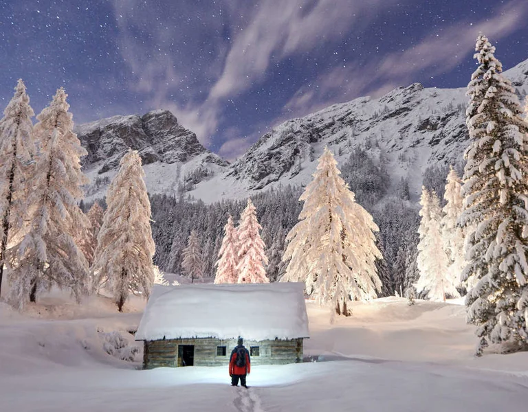 Snow-covered trees and cabin at night with a person standing in front of an illuminated cabin, and mountains in the background.