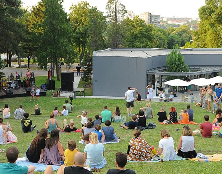Outdoor gathering with people sitting on grass enjoying a performance near a building.