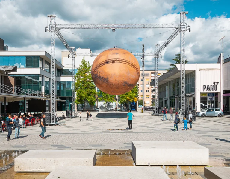 A public square with a large suspended sculpture of a planet, people walking, and cafes in the background.
