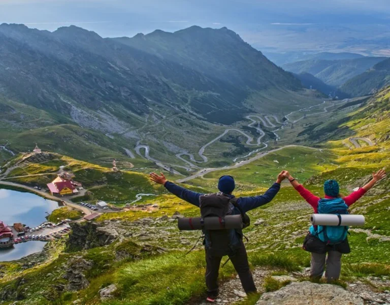 Two hikers with raised arms overlooking a mountain landscape with a winding road and lake.