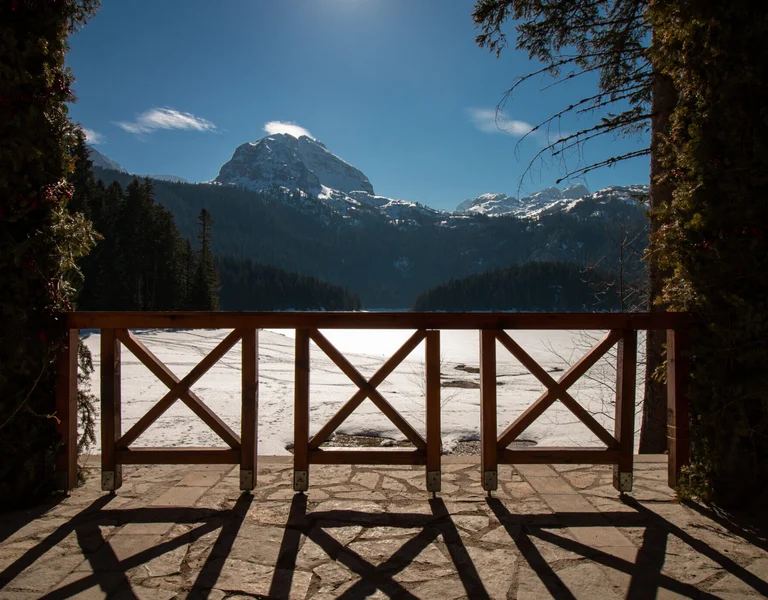 A serene view of snow-covered mountains and a frozen lake framed by the shadows of a rustic wooden balcony.