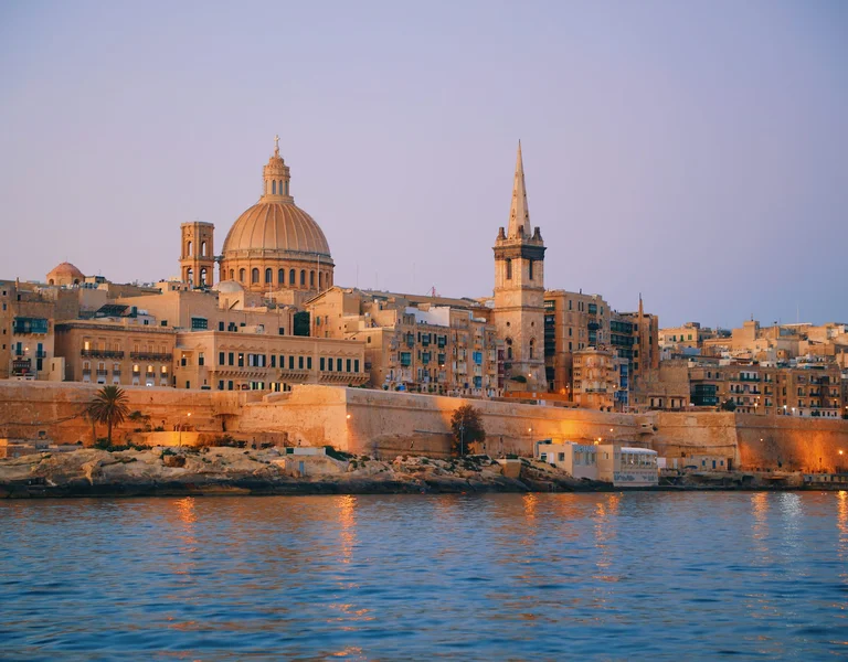 A view of Valletta, Malta, at sunset with its iconic domed church and spire illuminated by warm lights, reflected in the calm sea.