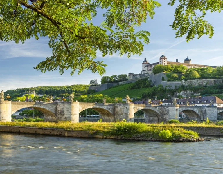 Historic stone bridge crossing a river with a hilltop castle and greenery under a clear sky.