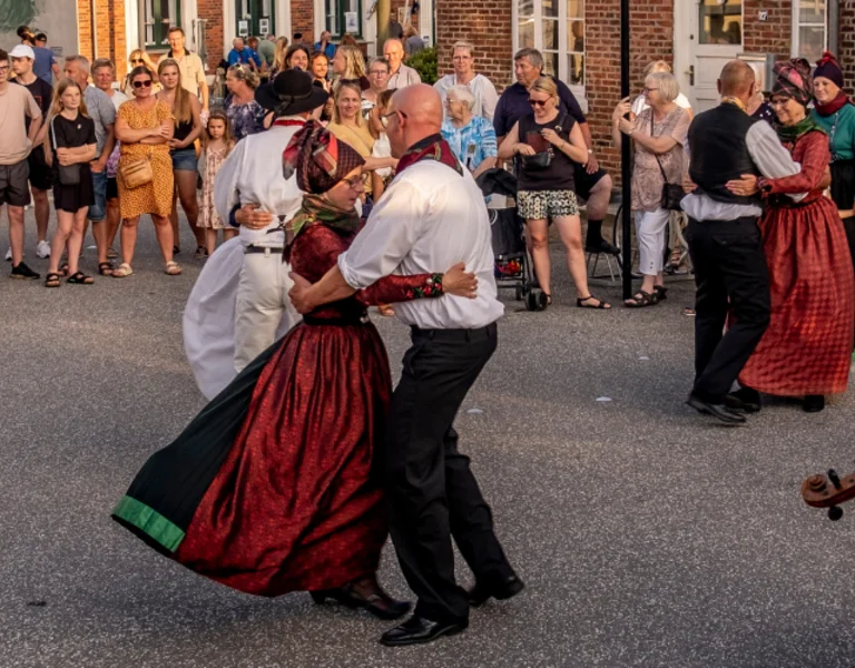 People in traditional attire dancing in a street with an onlooker playing a violin.
