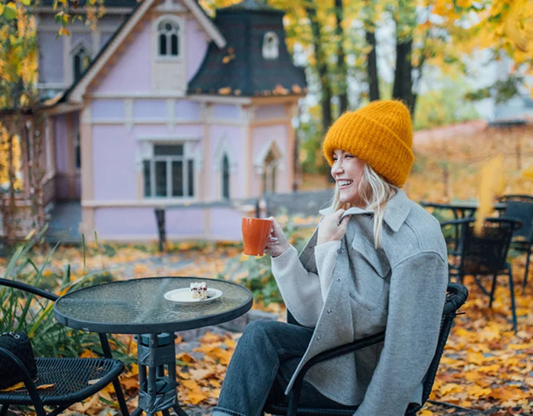 Person sitting at a cafe table with a drink during autumn, colorful house in background.