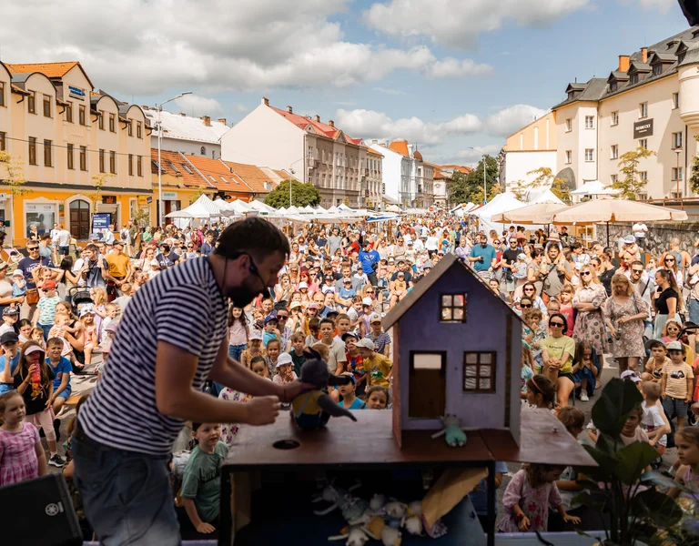 A man playing with a puppet, standing on a stage in front of a crowd in a colorful square.