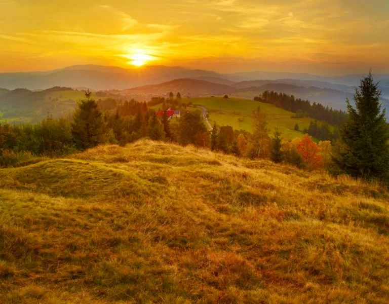 A sunset over rolling hills and forests, with a hint of mist and a small red-roofed house in the distance.
