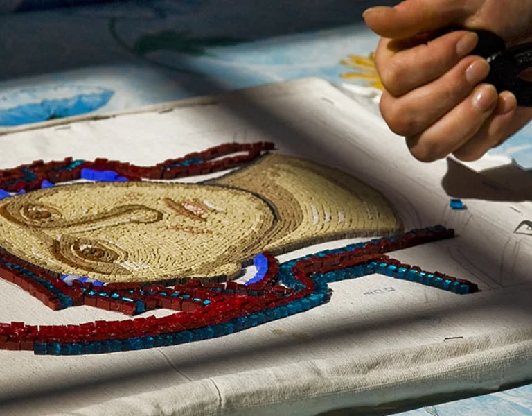 Close-up of hands assembling a mosaic of a face with colorful glass tiles on fabric, surrounded by tools and a patterned background.