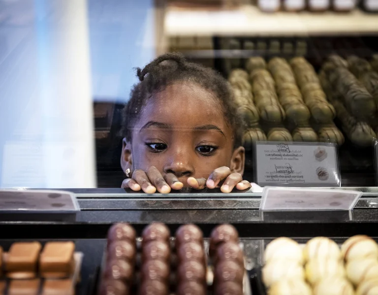 A kid with dark curly hair peers over the glass display case of a chocolate shop.