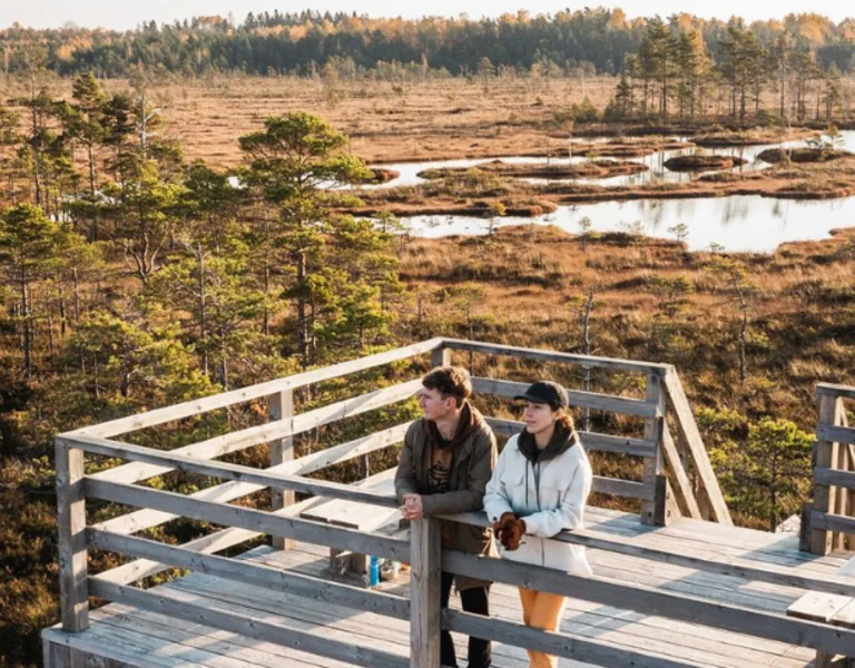 A couple stands on a wooden observation tower overlooking a scenic bog landscape with winding boardwalks, small lakes, and scattered pine trees in Estonia. The autumn colours create a warm atmosphere, with golden hues contrasting against the blue sky and water.