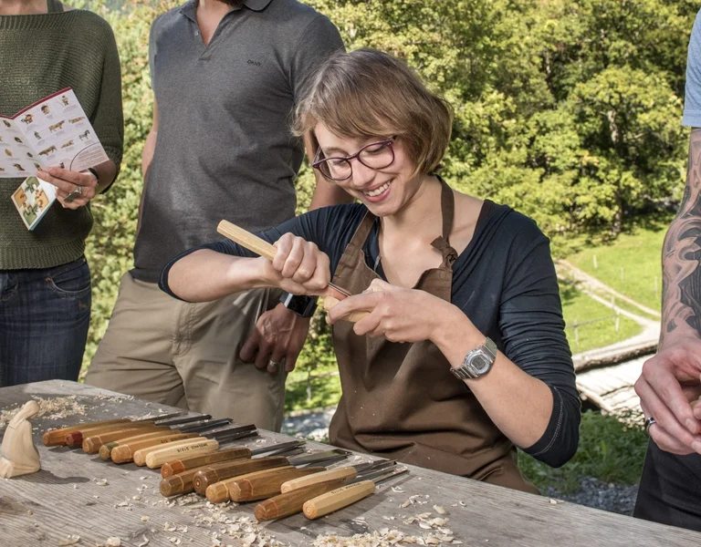 A group of four people is gathered around a wooden table, engaged in wood carving outdoors amidst scenic mountains and greenery.