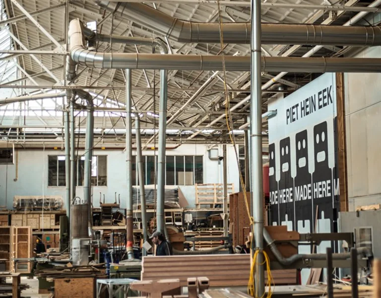 A spacious workshop filled with wooden materials, tools, and machinery, featuring a wall mural with "Piet Hein Eek" and figures.