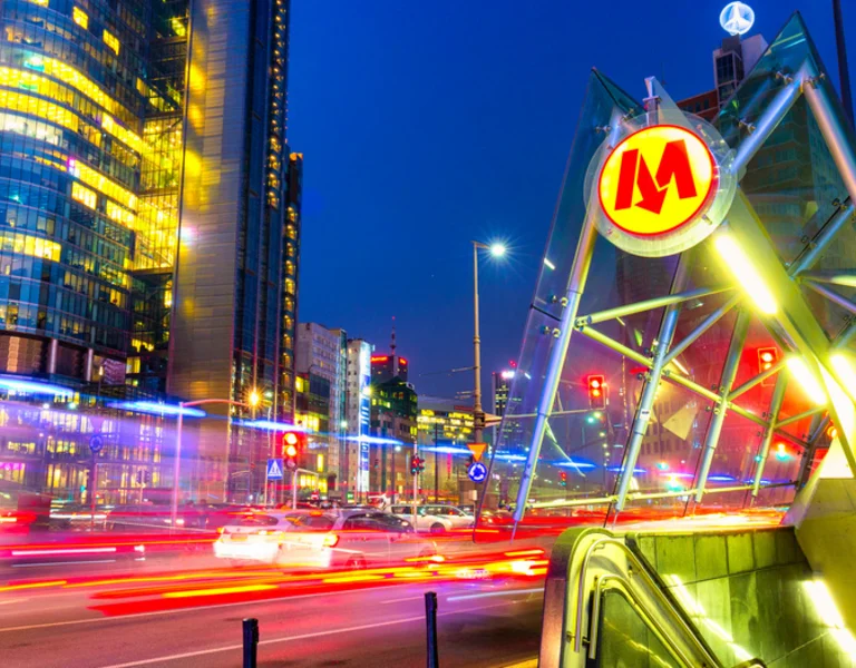 A modern metro station entrance in Warsaw illuminated at night, with skyscrapers and city traffic blurred in motion.