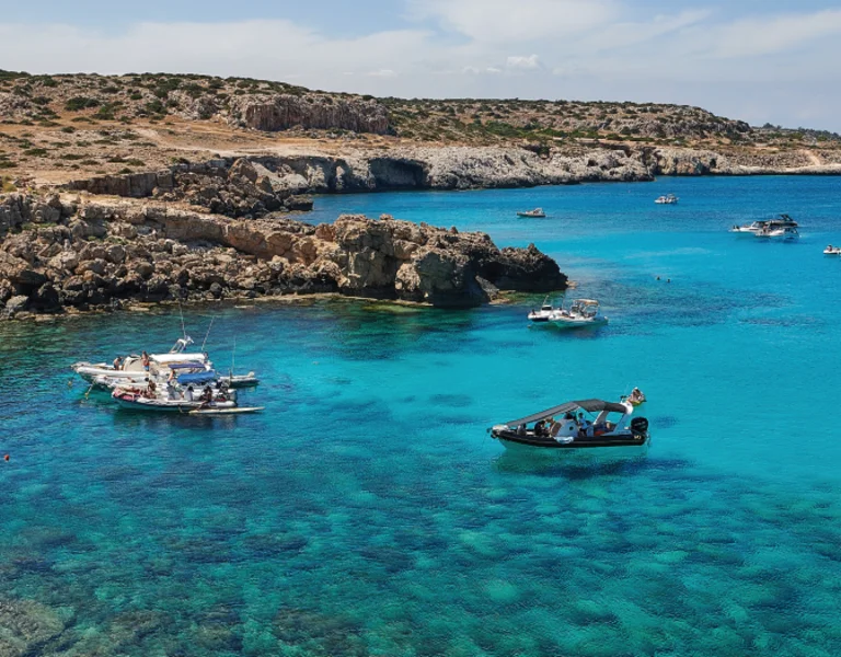 Turquoise waters and rocky cliffs of Cyprus, with several boats floating near the shore under a sunny sky.