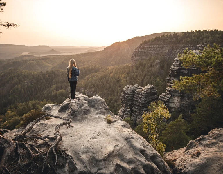 A girl stands on a rocky ledge, overlooking a vast forested valley at sunset, with mountains in the distance.
