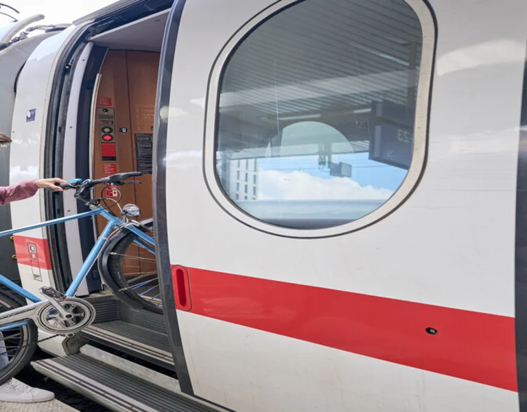 A person in a pink sweater is boarding a train while lifting a blue bicycle onto the carriage. The scene is set in the Frankfurt train station.
