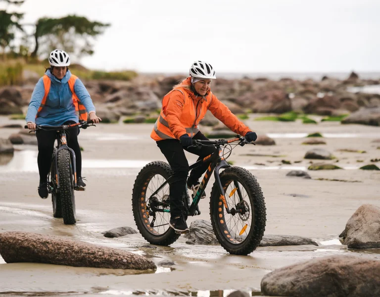 Two people wearing helmets and reflective jackets ride fat-tire bikes along a rocky and sandy beach. The lead cyclist, wearing an orange jacket, navigates carefully over the uneven terrain, while the second cyclist follows closely behind.