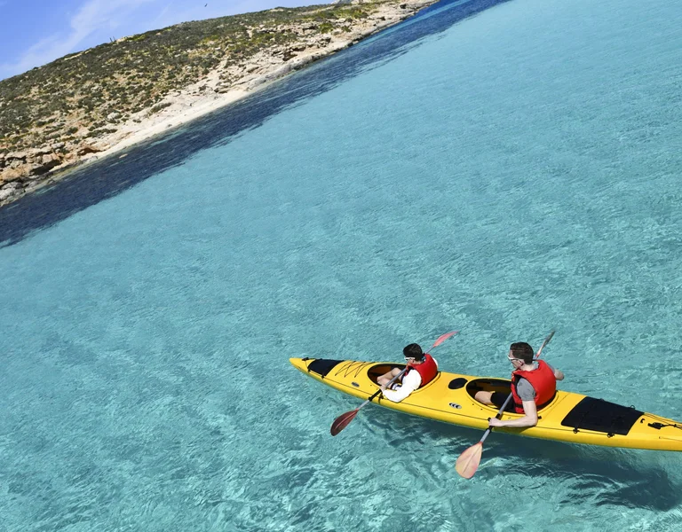 Two people wearing red life jackets paddle a yellow kayak through crystal-clear turquoise waters at the Blue Lagoon near a rocky coastline with green vegetation. The water is so clear that the sandy bottom is visible beneath the kayak.