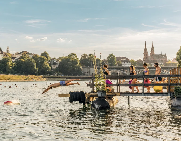 A summer scene by the river with swimmers and a diver, vibrant greenery, and historical buildings in the background.
