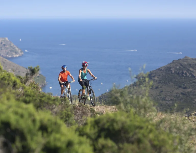 Two cyclists ride mountain bikes along a scenic coastal trail surrounded by lush greenery, with the deep blue sea and distant boats in the background.