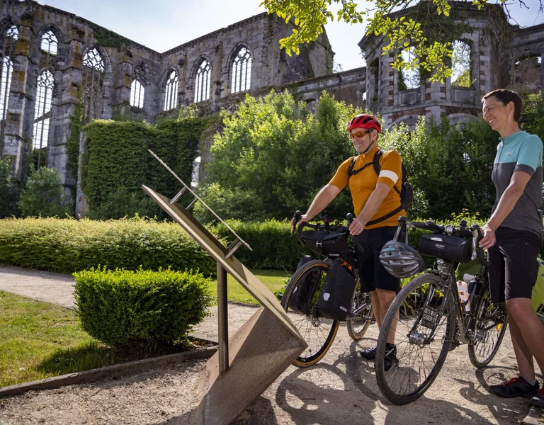 Two cyclists read an information board in front of the ivy-clad ruins of Aulne Abbey on a sunny day.