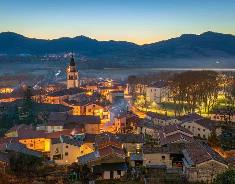 Twilight view of a tranquil village nestled amid mountains, featuring illuminated rooftops and a church steeple.