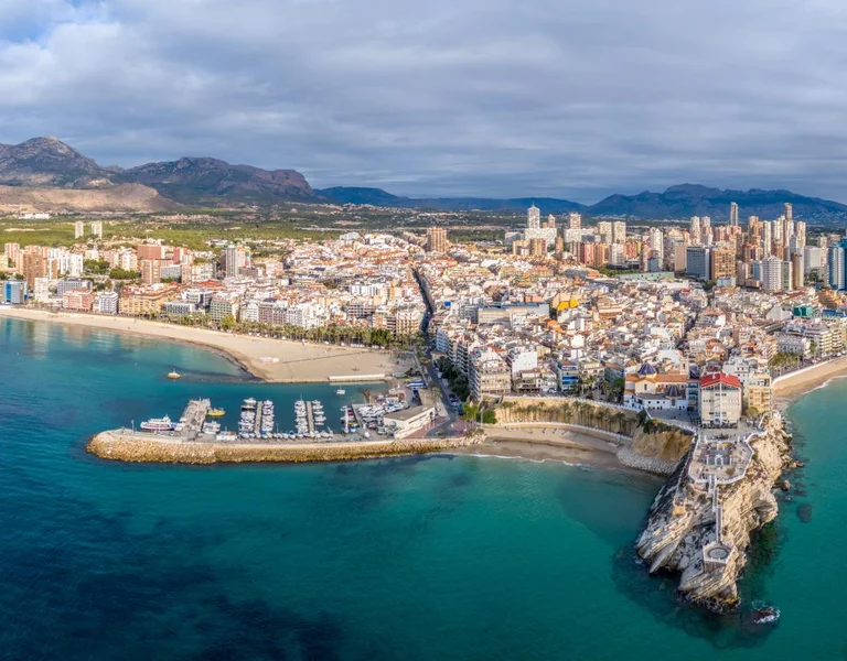 Aerial view of a coastal city with sandy beaches, marina, and modern buildings against a backdrop of mountains and cloudy skies.