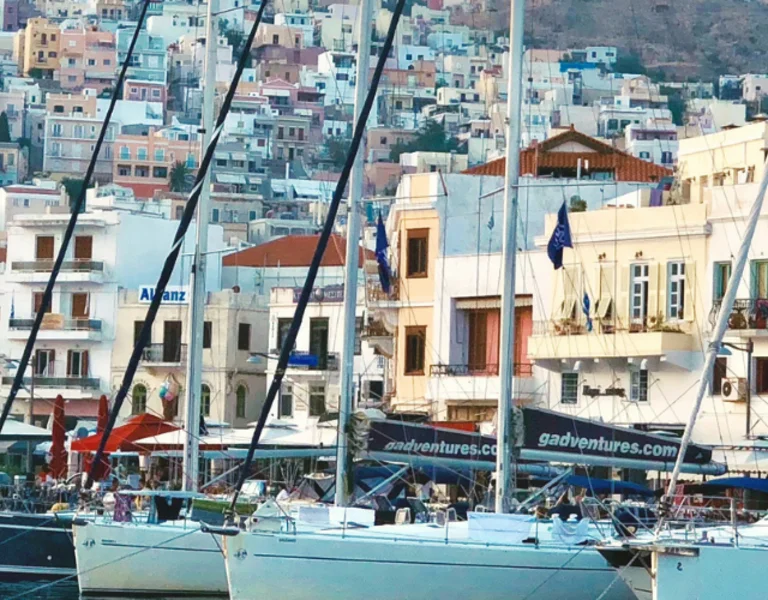 Sailboats docked at the vibrant harbor of Syros, Greece, with traditional houses climbing the hillside under a clear sky.