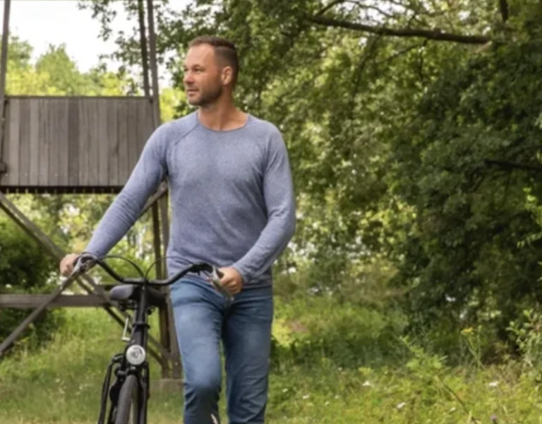 A couple walks along a grassy path, each holding a bicycle, with a wooden lookout tower and trees in the background.