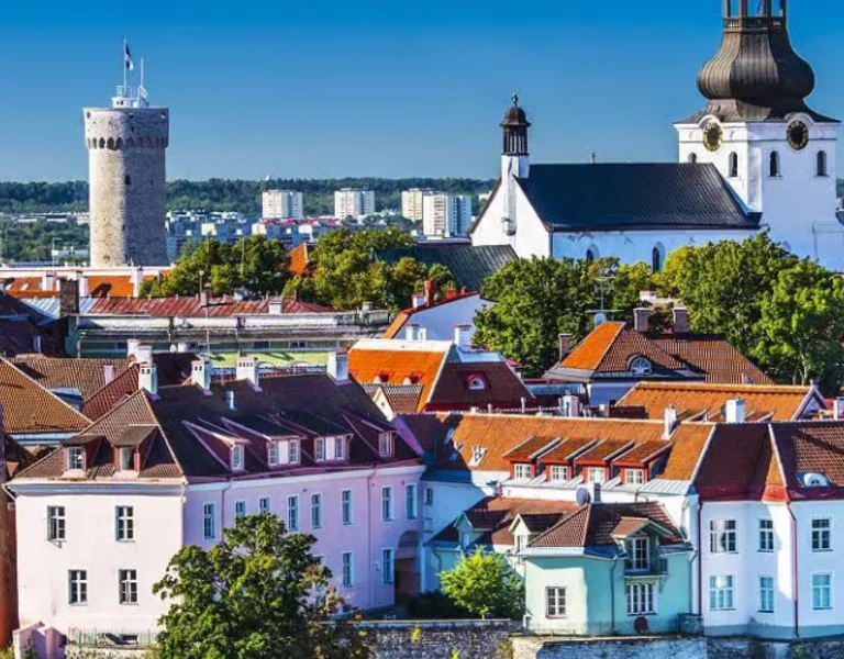 Aerial view of a historic European city at sunset, featuring a hilltop castle, red-roofed buildings, lush green parks, and a river with a docked boat.