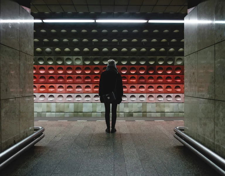 A person stands silhouetted in a subway corridor, facing a colorful, textured wall of red and green panels under dim lighting.