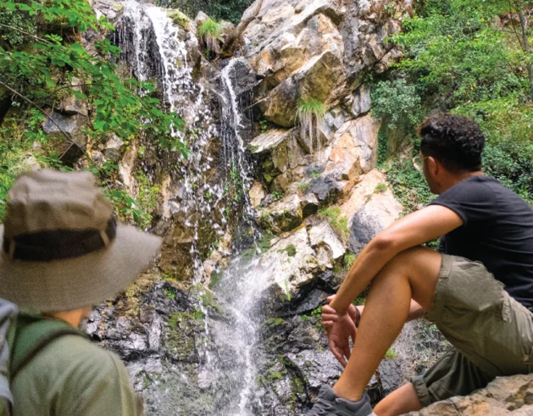 Two hikers sitting near a cascading waterfall in a lush forest, surrounded by vibrant greenery and rocky terrain.