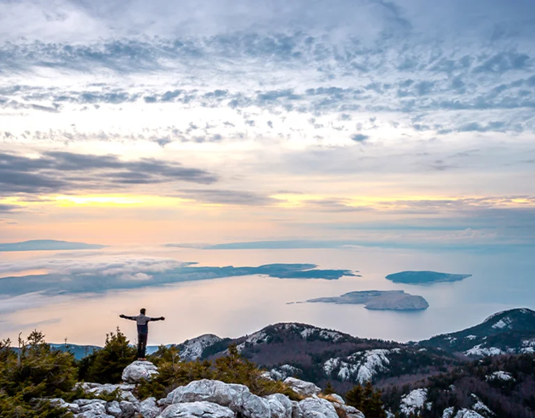 A person stands on a rocky peak, arms outstretched, overlooking serene islands and a misty sea under a colorful sunset sky.