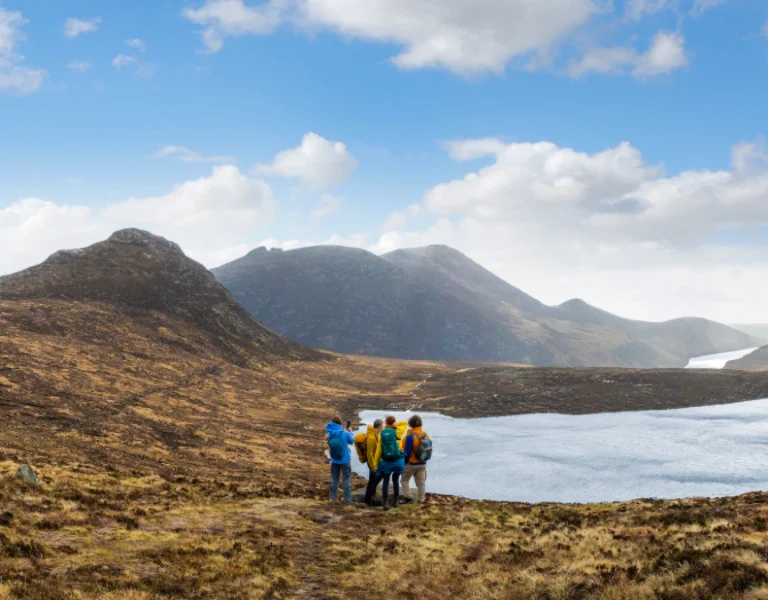 A group of hikers stands on a grassy hillside, overlooking a serene lake surrounded by mountains under a partly cloudy sky.