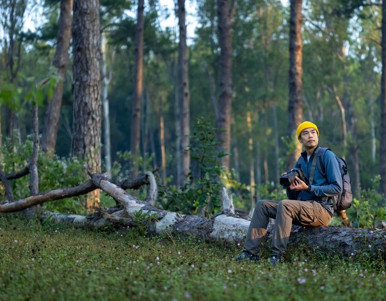 A man wearing a yellow beanie and outdoor gear sits on a fallen tree trunk in a forest, holding a camera and gazing into the distance, surrounded by tall pine trees and lush greenery.