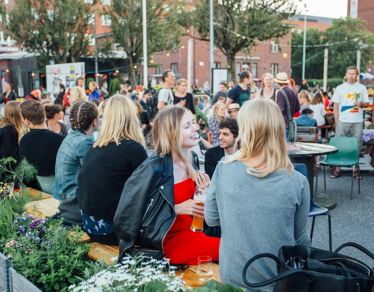 A vibrant outdoor gathering with people socializing, enjoying drinks, and surrounded by greenery and festive decor.