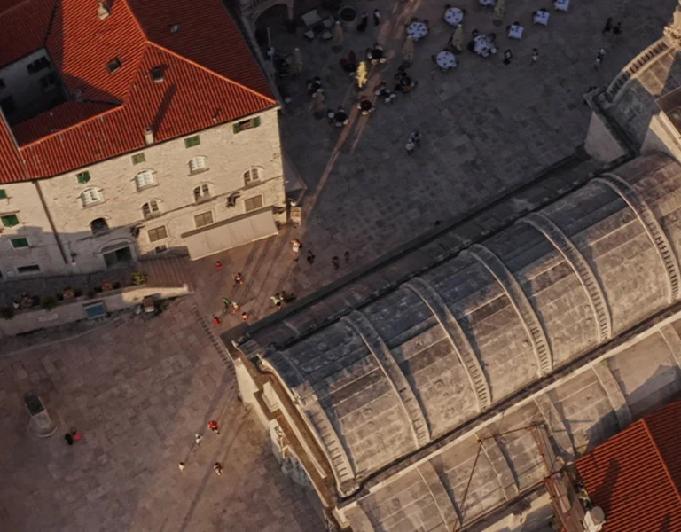 Aerial view of a square surrounded by buildings with red roofs and a prominent church with a dome, bustling with people and outdoor seating.
