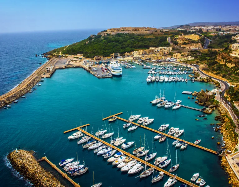 Aerial view of a vibrant marina with numerous yachts docked, surrounded by turquoise waters and lush green hills in the background.