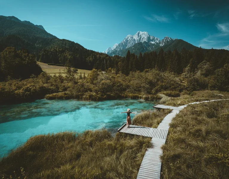 A person stands with arms outstretched on a wooden boardwalk beside a crystal-clear turquoise pond, surrounded by grassy wetlands and dense forest, with dramatic snow-capped mountains rising in the background under a vivid blue sky.