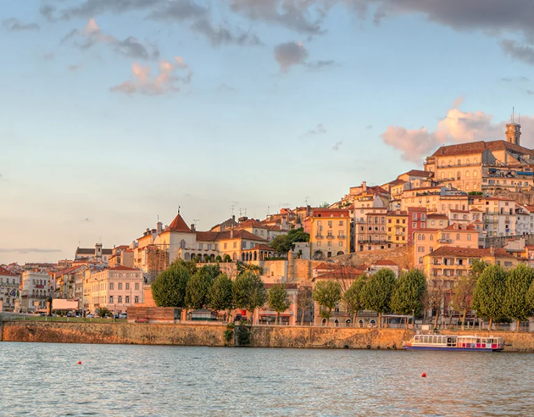 Panoramic view of a riverside city at sunset, showcasing picturesque buildings on a hill and a bridge crossing the water.