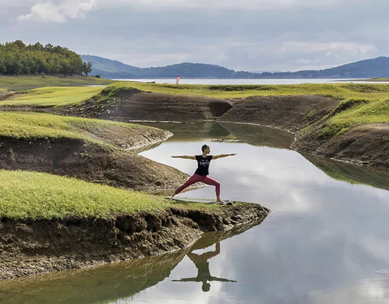 Person practising yoga by a reflective stream in the green, hilly landscape of Lake Plastiras, Thessaly, Greece.