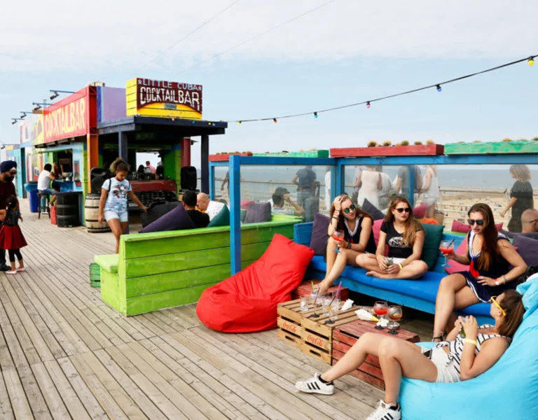 Group of friends relaxing with drinks at a colourful beach cocktail bar on a sunny boardwalk in Scheveningen, Netherlands.