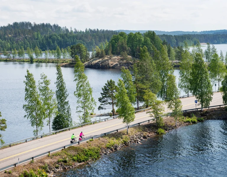 Two cyclists riding along a scenic lakeside road surrounded by forest in Finland's Lakeland region.