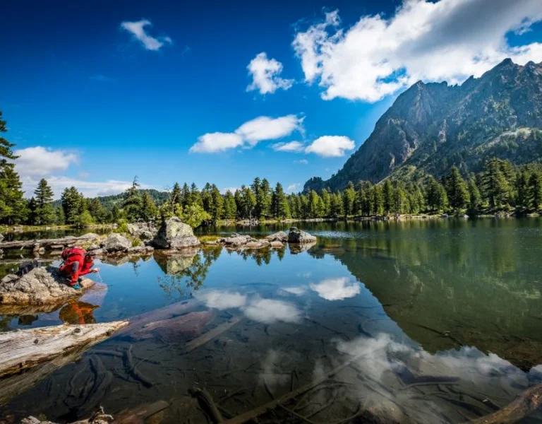 Hiker resting by the serene Hridsko Lake, surrounded by pine trees and mountains under a bright blue sky in Montenegro.