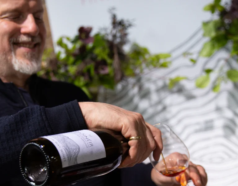 A man pours a glass of dark liquid from a bottle, surrounded by greenery and wooden shelves in a cozy setting.