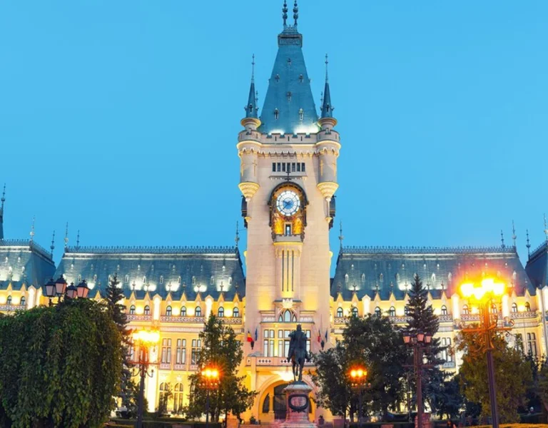 Illuminated palace with intricate architecture and a clock tower, surrounded by lush greenery and a peaceful evening sky.